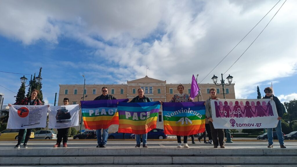 7 άτομα στην πλατεία Συντάγματος με φόντο το κτίριο της Βουλής των Ελλήνων κρατούν πολύχρωμα πανό αλληλεγγύης στους αντιρρησίες συνείδησης του Object War Campaign με κείμενα "Peace", "Pace", "Nonviolenza", "το μωβ (tomov.gr)"